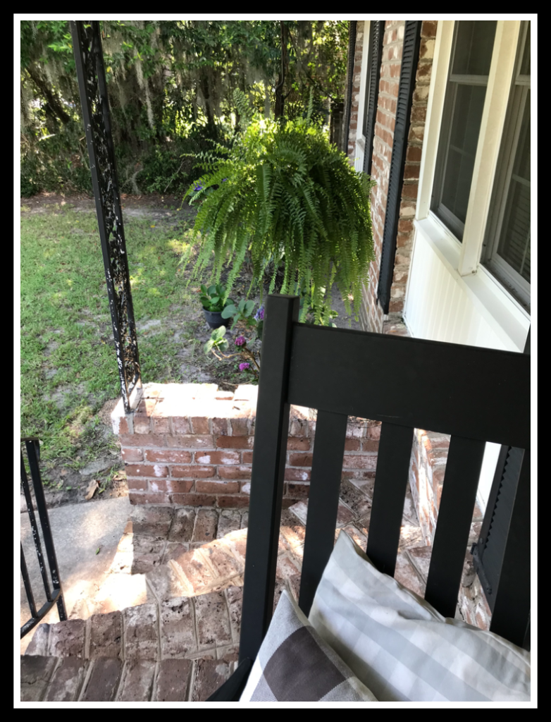 Black rocking chair on cozy front porch with green hanging fern in the background