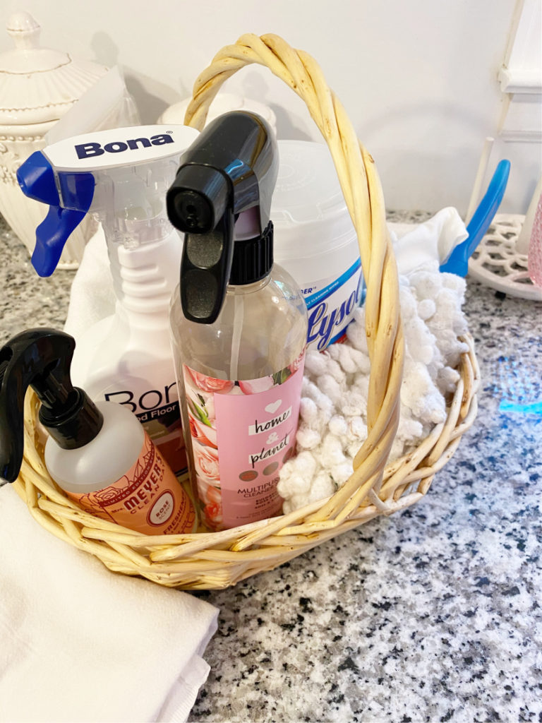 Basket of cleaning supplies with duster and white towel beside it on black and white granite counter