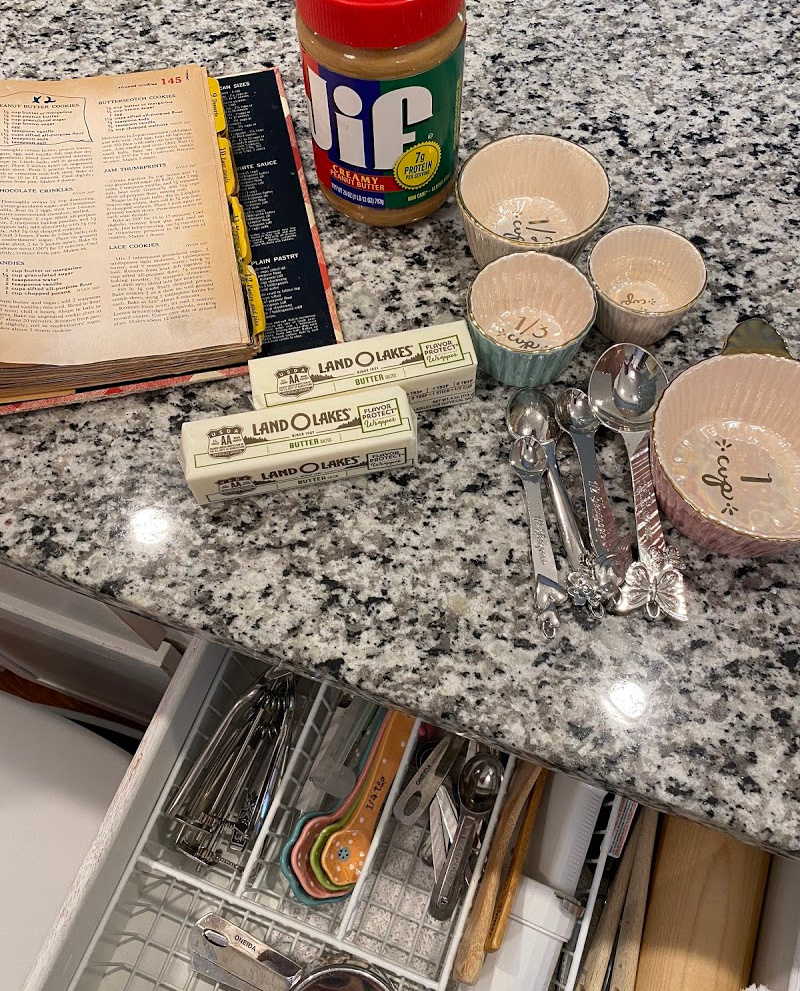 Cook book open with butter, measuring spoons, measuring cups, and open utensil drawer